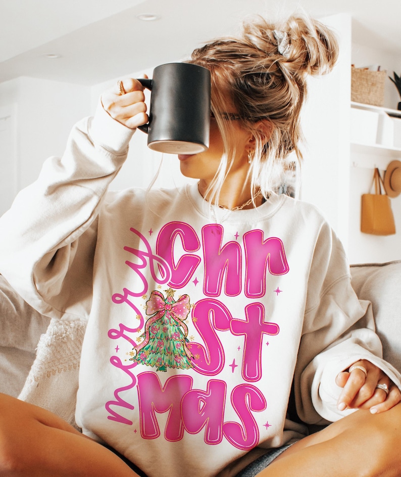 May include: Cream-colored sweatshirt with the words "Merry Christmas" in pink, featuring a Christmas tree graphic. The tree is green with a pink bow and gold accents. The sweatshirt is a casual, festive garment.