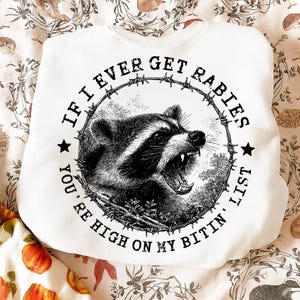 May include: White sweatshirt with a graphic of a snarling raccoon inside a barbed wire circle. The text around the raccoon reads "IF I EVER GET RABIES YOU'RE HIGH ON MY BITIN' LIST."