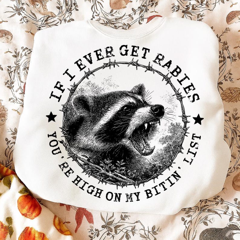 May include: White sweatshirt with a graphic of a snarling raccoon inside a barbed wire circle. The text around the raccoon reads "IF I EVER GET RABIES YOU'RE HIGH ON MY BITIN' LIST."