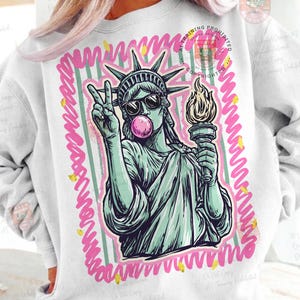 Puede incluir: Sudadera blanca con un gr&aacute;fico de la Estatua de la Libertad con gafas de sol y soplando una burbuja de chicle. La Estatua de la Libertad sostiene una antorcha y hace el signo de la paz. El gr&aacute;fico est&aacute; enmarcado por un borde rosa y amarillo.