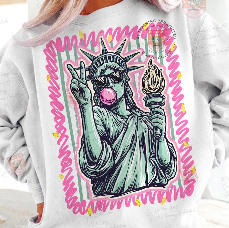 Puede incluir: Sudadera blanca con un gr&aacute;fico de la Estatua de la Libertad con gafas de sol y soplando una burbuja de chicle. La Estatua de la Libertad sostiene una antorcha y hace el signo de la paz. El gr&aacute;fico est&aacute; enmarcado por un borde rosa y amarillo.