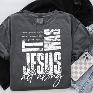May include: Dark gray t-shirt with the text "IT WAS JESUS ALL ALONG" in white. The shirt also features the words "faith, grace, truth, saved, amen, love, holy, peace, spirit." A phone with a checkered case and blue jeans are also visible.