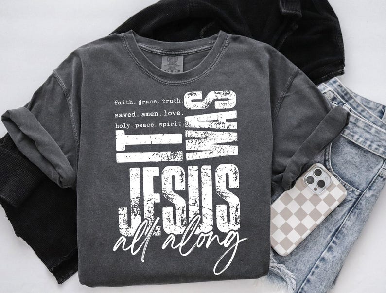 May include: Dark gray t-shirt with the text "IT WAS JESUS ALL ALONG" in white. The shirt also features the words "faith, grace, truth, saved, amen, love, holy, peace, spirit." A phone with a checkered case and blue jeans are also visible.