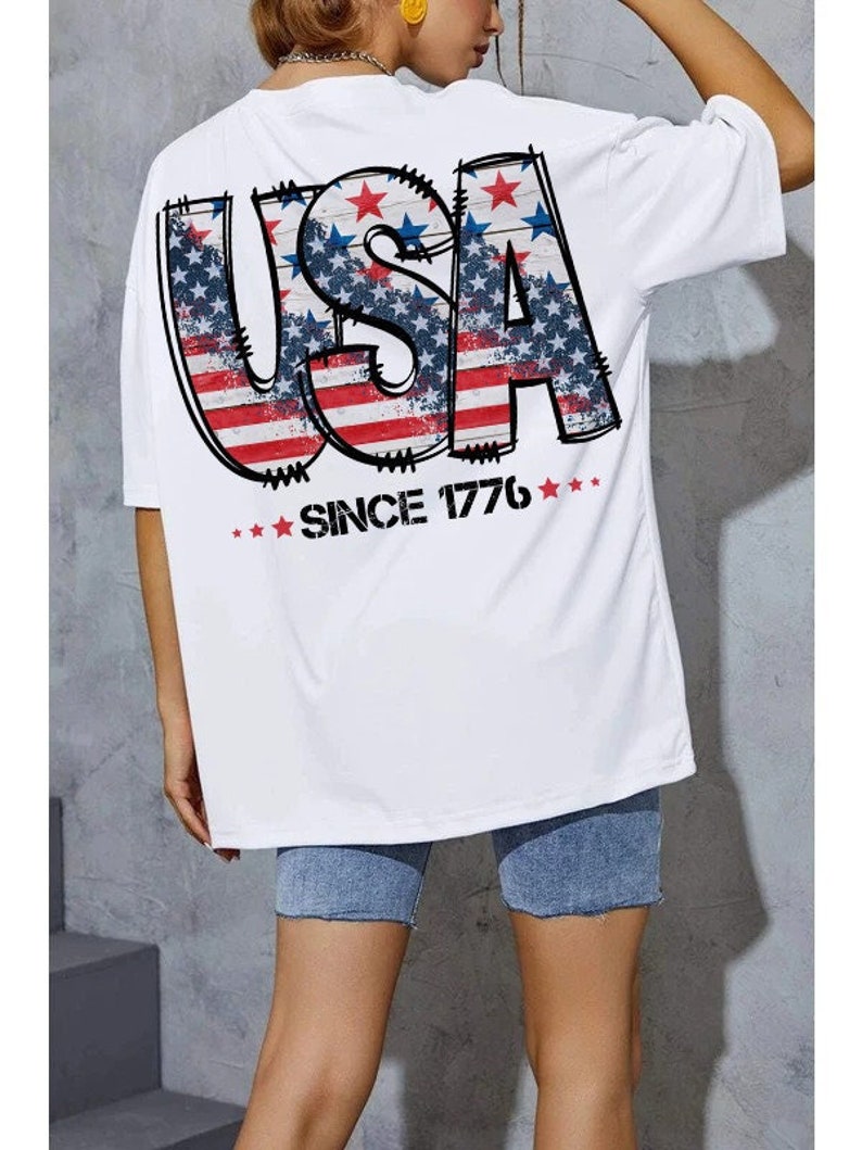 May include: White t-shirt with a graphic of the word "USA" in red, white, and blue with stars and stripes. The text "Since 1776" is printed below the graphic.