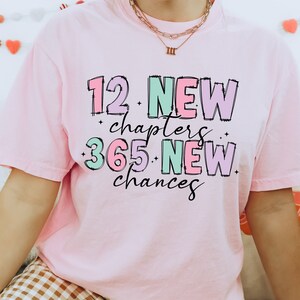 K&ouml;nnte beinhalten: Rosa T-Shirt mit dem Text "12 New chapters 365 New chances" in bunten, umrandeten Buchstaben.