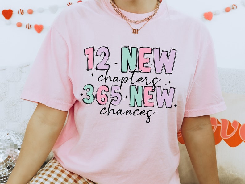 K&ouml;nnte beinhalten: Rosa T-Shirt mit dem Text "12 New chapters 365 New chances" in bunten, umrandeten Buchstaben.