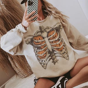 May include: Beige sweatshirt featuring a skeletal rib cage design with jack-o'-lanterns inside. The sweatshirt has long sleeves and a crew neck. A white baseball cap with "Malibu Cowboy" text is also visible.