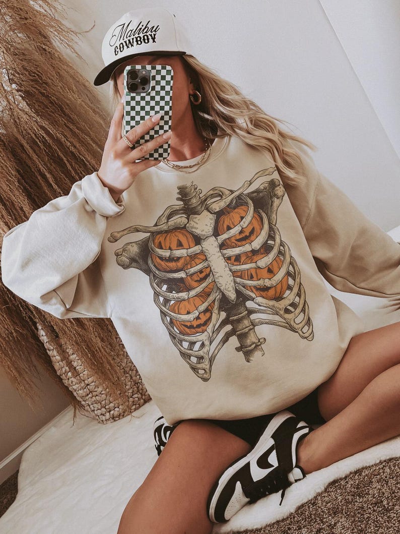 May include: Beige sweatshirt featuring a skeletal rib cage design with jack-o'-lanterns inside. The sweatshirt has long sleeves and a crew neck. A white baseball cap with "Malibu Cowboy" text is also visible.