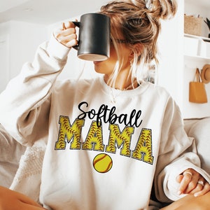 May include: White sweatshirt with the text "Softball Mama" in black lettering. The letters are outlined in yellow and have a red stitching design. A yellow softball with red stitching is below the text.