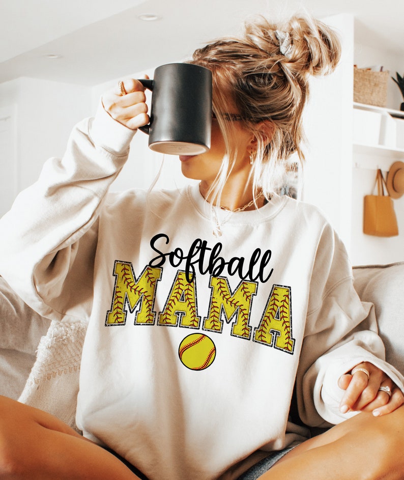 May include: White sweatshirt with the text "Softball Mama" in black lettering. The letters are outlined in yellow and have a red stitching design. A yellow softball with red stitching is below the text.