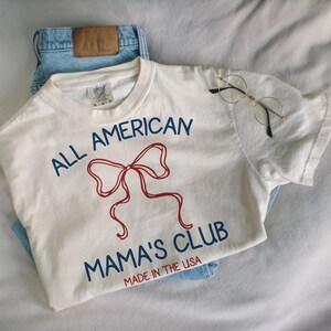 May include: An off-white t-shirt with the text "ALL AMERICAN MAMA'S CLUB" in blue and a red bow graphic. The shirt is paired with light blue jeans and round-framed glasses.