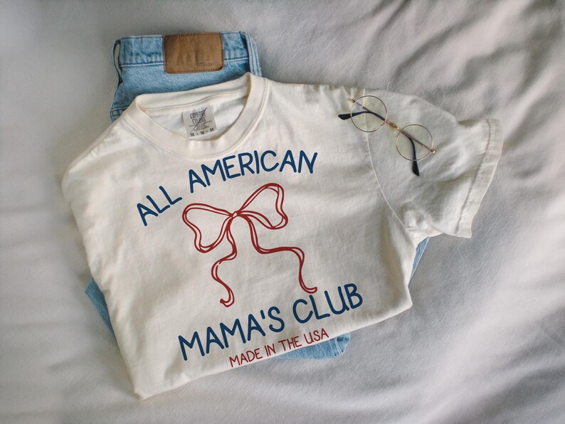 May include: An off-white t-shirt with the text "ALL AMERICAN MAMA'S CLUB" in blue and a red bow graphic. The shirt is paired with light blue jeans and round-framed glasses.
