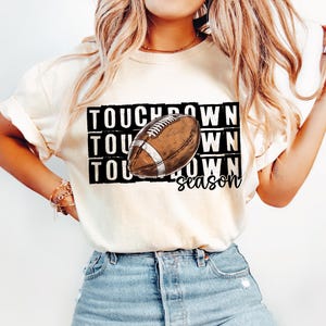 Op de afbeelding: Een wit T-shirt met de tekst "Touchdown Season" in zwart gedrukt met een bruine en witte voetbalafbeelding.