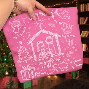 K&ouml;nnte beinhalten: Ein pinkfarbener Kissenbezug mit wei&szlig;en Illustrationen und Text zum Thema Weihnachten. Das Design umfasst einen Engel, einen Weihnachtsbaum, eine Krippe, ein Geschenk, ein Schaf und die S&auml;tze "Joy to the World" und "Peace on Earth."