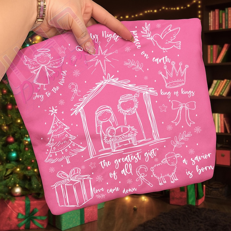 K&ouml;nnte beinhalten: Ein pinkfarbener Kissenbezug mit wei&szlig;en Illustrationen und Text zum Thema Weihnachten. Das Design umfasst einen Engel, einen Weihnachtsbaum, eine Krippe, ein Geschenk, ein Schaf und die S&auml;tze "Joy to the World" und "Peace on Earth."