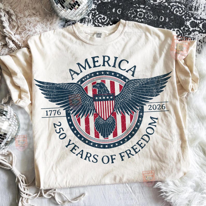 May include: Cream-colored t-shirt featuring a patriotic design. The graphic includes the word "AMERICA" in a circular banner, an eagle with outstretched wings, and the text "250 YEARS OF FREEDOM" with the years 1776 and 2026.