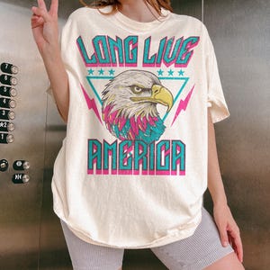 May include: A white t-shirt with a graphic of an eagle in a triangle with the text "Long Live America" in turquoise and pink.