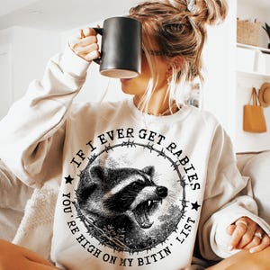 May include: Cream-colored sweatshirt with a graphic of a raccoon inside a barbed wire circle. The text reads "If I Ever Get Rabies, You're High On My Bitin' List." The raccoon is black and white with an open mouth.