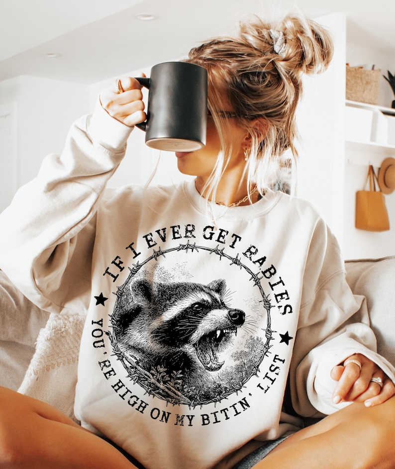 May include: Cream-colored sweatshirt with a graphic of a raccoon inside a barbed wire circle. The text reads "If I Ever Get Rabies, You're High On My Bitin' List." The raccoon is black and white with an open mouth.