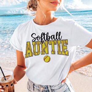 May include: White t-shirt with the text "Softball Auntie" in black lettering with a yellow softball graphic. The text is outlined with a yellow stitching design.