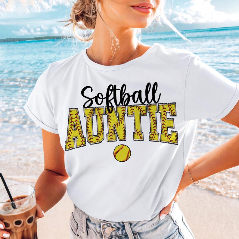 May include: White t-shirt with the text "Softball Auntie" in black lettering with a yellow softball graphic. The text is outlined with a yellow stitching design.