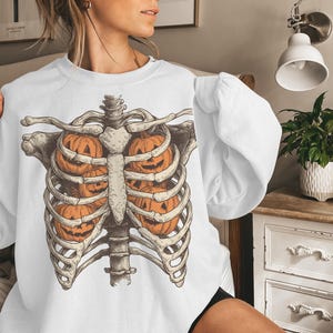 May include: White sweatshirt featuring a Halloween-themed design. The graphic depicts a skeletal rib cage with orange jack-o'-lanterns in place of the internal organs. The design is detailed and has a vintage aesthetic.