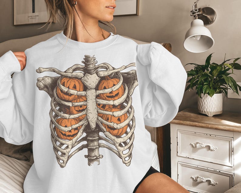 May include: White sweatshirt featuring a Halloween-themed design. The graphic depicts a skeletal rib cage with orange jack-o'-lanterns in place of the internal organs. The design is detailed and has a vintage aesthetic.
