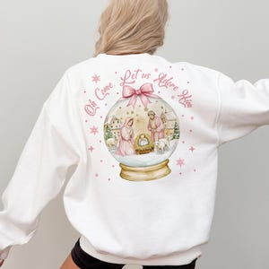 K&ouml;nnte beinhalten: Wei&szlig;er Pullover mit einem rosa und goldenen Schneekugel-Design, das eine Weihnachtskrippe zeigt. Der Text "Oh Come, Let us Adore Him" ist in rosa auf der R&uuml;ckseite des Pullovers gedruckt.