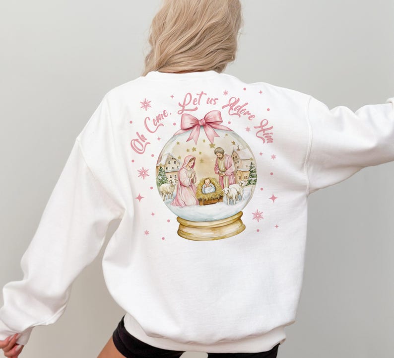 K&ouml;nnte beinhalten: Wei&szlig;er Pullover mit einem rosa und goldenen Schneekugel-Design, das eine Weihnachtskrippe zeigt. Der Text "Oh Come, Let us Adore Him" ist in rosa auf der R&uuml;ckseite des Pullovers gedruckt.