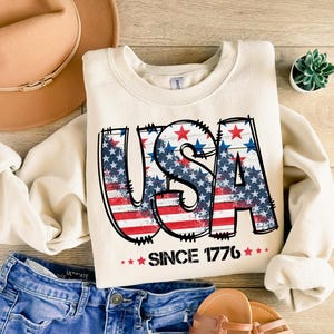 May include: Cream-colored sweatshirt with the word "USA" in a distressed design featuring the American flag. The text "SINCE 1776" is below the word "USA". The sweatshirt is paired with blue denim shorts, a tan hat, and sandals.