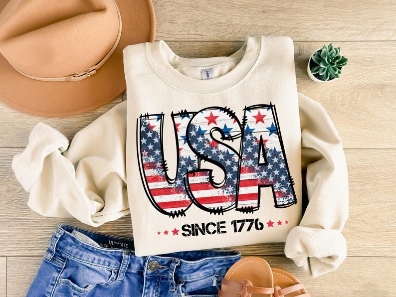 May include: Cream-colored sweatshirt with the word "USA" in a distressed design featuring the American flag. The text "SINCE 1776" is below the word "USA". The sweatshirt is paired with blue denim shorts, a tan hat, and sandals.