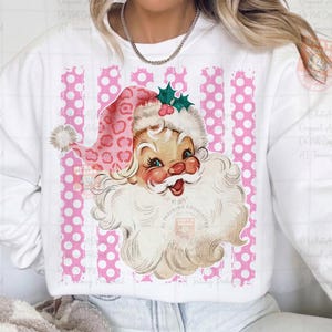 Pode incluir: Uma camisola branca com um gr&aacute;fico de um Pai Natal de estilo vintage a usar um gorro com estampado de leopardo cor-de-rosa e um ramo de azevinho verde. O fundo do gr&aacute;fico consiste em riscas verticais cor-de-rosa com bolas brancas.