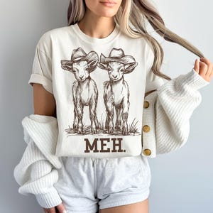 May include: A white t-shirt with a brown illustration of two goats wearing cowboy hats. The text "MEH." is printed below the goats.