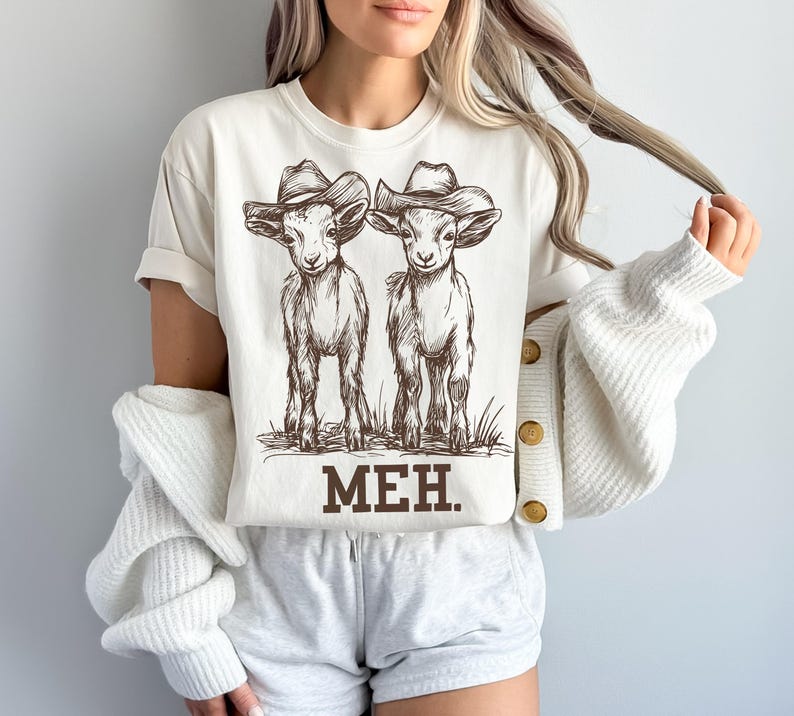 May include: A white t-shirt with a brown illustration of two goats wearing cowboy hats. The text "MEH." is printed below the goats.