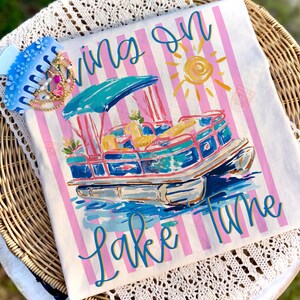 May include: White t-shirt with pink and white stripes, featuring a colorful illustration of a pontoon boat on water. The shirt has the words "Living on Lake Time" in blue script. A blue hair clip with gold and pink accents is on the side.