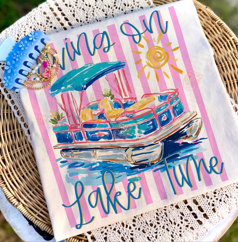 May include: White t-shirt with pink and white stripes, featuring a colorful illustration of a pontoon boat on water. The shirt has the words "Living on Lake Time" in blue script. A blue hair clip with gold and pink accents is on the side.