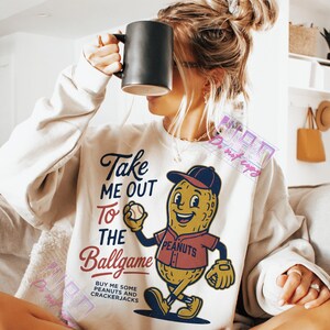 May include: Cream-colored sweatshirt with a baseball-themed graphic. The graphic features a smiling peanut character in a baseball uniform, holding a baseball. Text on the shirt reads "Take Me Out To The Ballgame" and "Buy Me Some Peanuts and Crackerjacks."