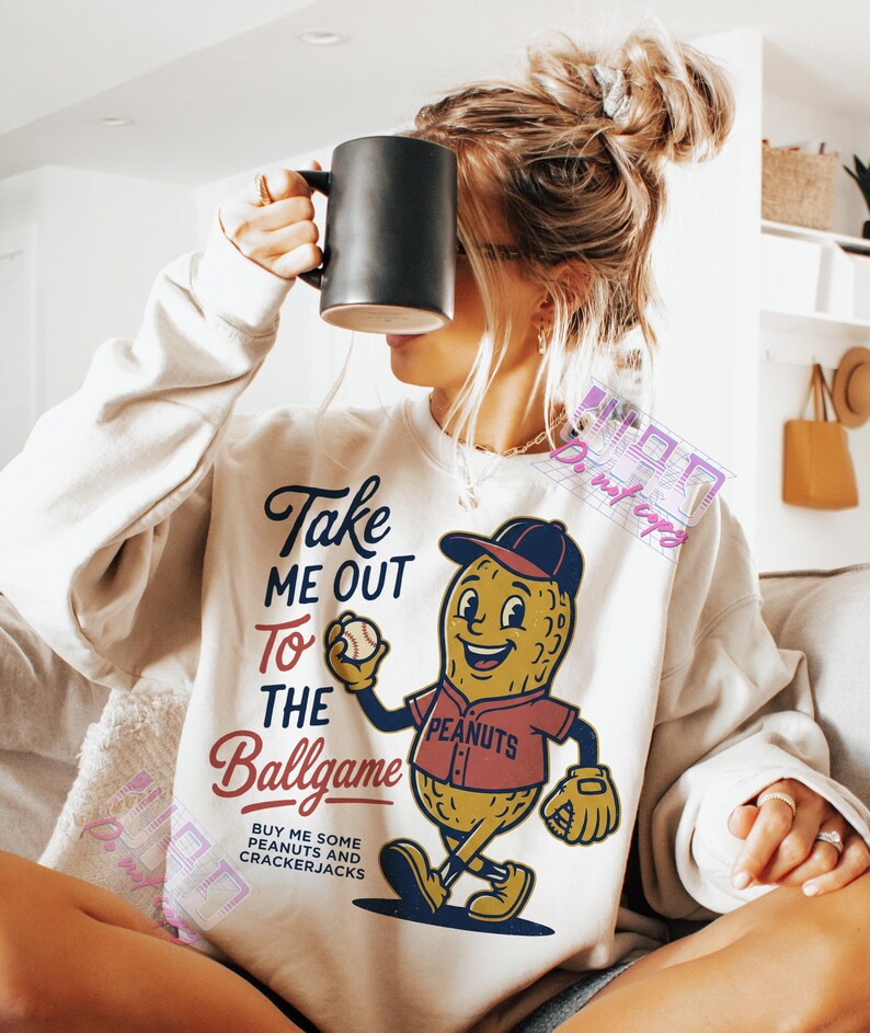 May include: Cream-colored sweatshirt with a baseball-themed graphic. The graphic features a smiling peanut character in a baseball uniform, holding a baseball. Text on the shirt reads "Take Me Out To The Ballgame" and "Buy Me Some Peanuts and Crackerjacks."