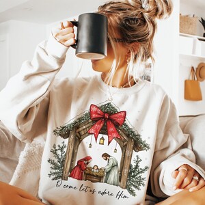 May include: Cream-colored sweatshirt featuring a Nativity scene with a red bow, evergreen accents, and the text "O come let us adore Him." The design includes a stable, figures, and a baby. The person in the image is holding a black mug.
