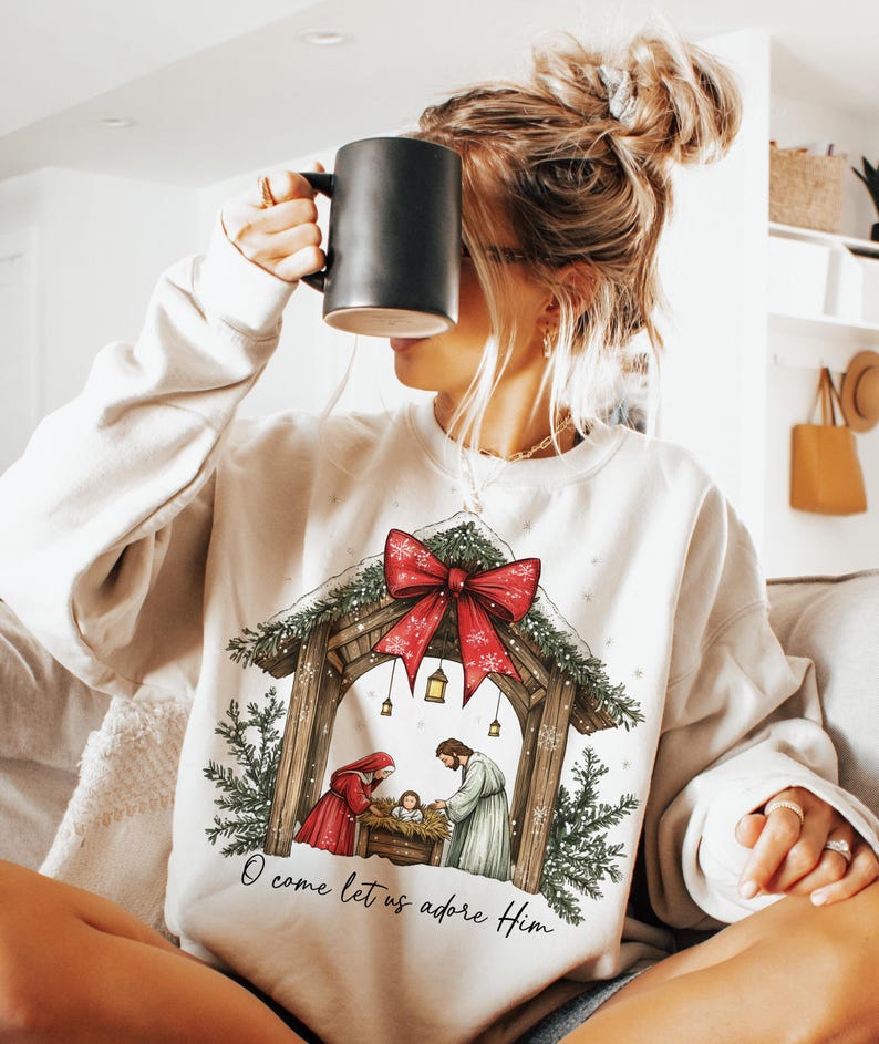 May include: Cream-colored sweatshirt featuring a Nativity scene with a red bow, evergreen accents, and the text "O come let us adore Him." The design includes a stable, figures, and a baby. The person in the image is holding a black mug.