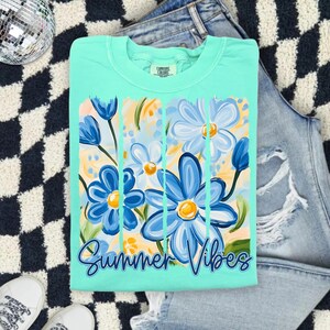 May include: A turquoise blue t-shirt with a graphic of blue and yellow flowers and the text "Summer Vibes".