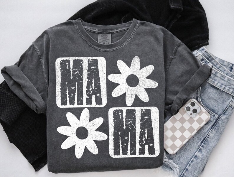 Peut inclure: Un t-shirt gris fonc&eacute; avec un motif graphique blanc. Le graphique pr&eacute;sente le mot "mama" dans une police de caract&egrave;res us&eacute;e, entour&eacute; de marguerites blanches stylis&eacute;es.