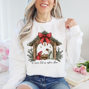 May include: White sweatshirt featuring a Christmas Nativity scene with a red bow, evergreen accents, and the text "O come let us adore Him." The illustration depicts Mary, Joseph, and baby Jesus in a manger. A festive holiday-themed top.