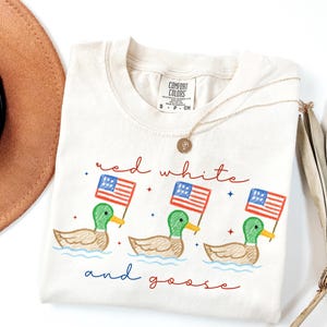 May include: Cream-colored Comfort Colors t-shirt with a graphic of three ducks holding American flags. The text "red white and goose" is written in red and blue script. A gold necklace is draped on the shirt.