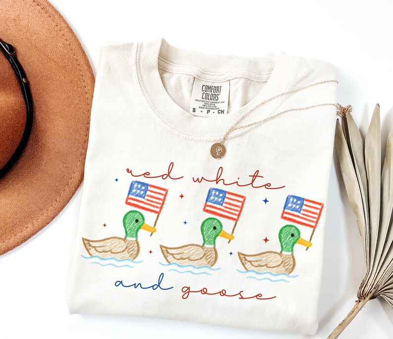 May include: Cream-colored Comfort Colors t-shirt with a graphic of three ducks holding American flags. The text "red white and goose" is written in red and blue script. A gold necklace is draped on the shirt.