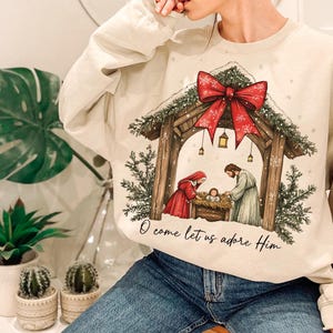 May include: Cream-colored sweatshirt featuring a Christmas nativity scene with a red bow and the text "O come let us adore Him." The graphic depicts Mary, Joseph, and baby Jesus in a manger. The sweatshirt is a cozy, festive garment.