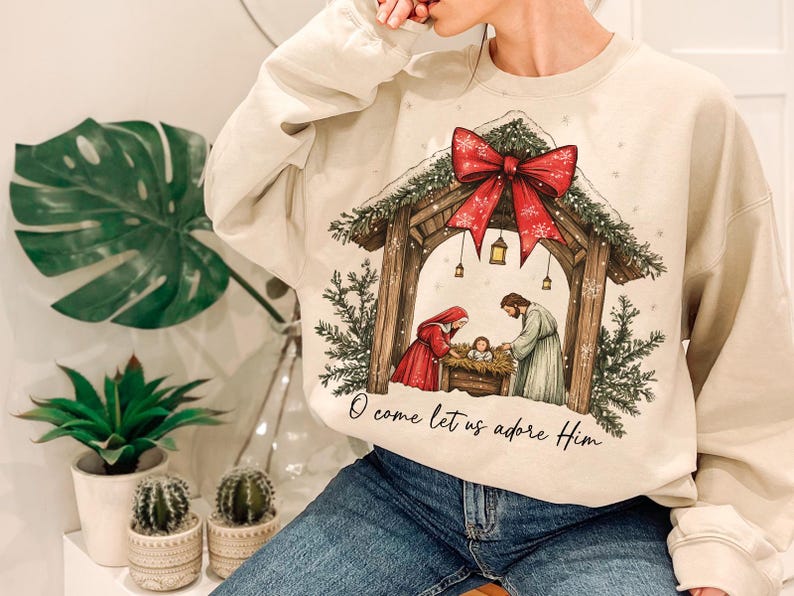 May include: Cream-colored sweatshirt featuring a Christmas nativity scene with a red bow and the text "O come let us adore Him." The graphic depicts Mary, Joseph, and baby Jesus in a manger. The sweatshirt is a cozy, festive garment.