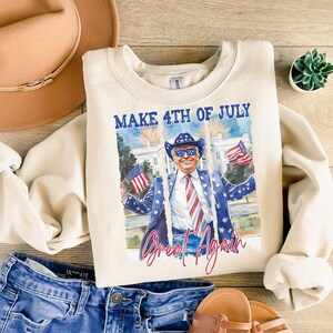 May include: A cream-colored sweatshirt with a graphic print of a man wearing sunglasses and a red, white, and blue tie. The text "Make 4th of July Great Again" is printed on the sweatshirt.