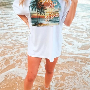 May include: A white t-shirt with a graphic print of a sunset over the ocean with palm trees and the text "salt water and sun shine".