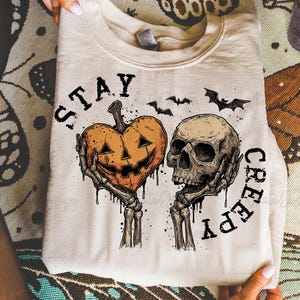 Puede incluir: Sudadera beige con dise&ntilde;o de Halloween. El gr&aacute;fico incluye un coraz&oacute;n de calabaza y una calavera sostenidos por manos esquel&eacute;ticas, con las palabras "STAY CREEPY" y siluetas de murci&eacute;lagos. El dise&ntilde;o es en negro y naranja.